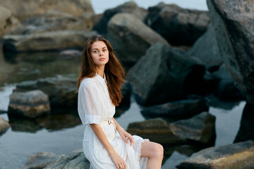 Serene woman in white dress sitting on rocky shore, gazing out at tranquil water