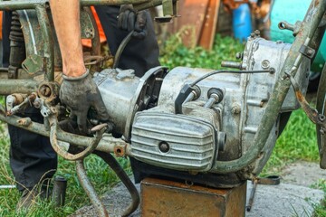worker with mittened hands removes one industrial iron dirty old two-cylinder oily broken engine from  rusty frame of green retro motorcycle during daytime on street