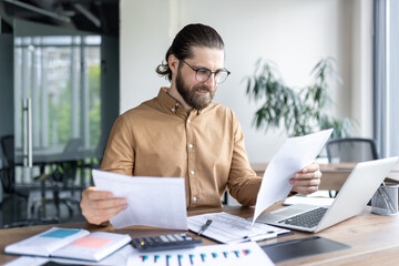 Obraz premium Smiling businessman wearing eyeglasses analyzing reports at desk with laptop, papers, and calculator. Professional in modern office environment, focusing on data and strategy for business growth.