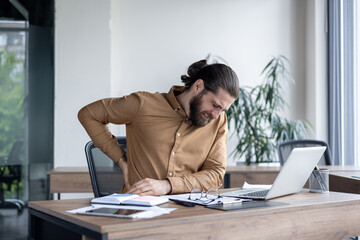 Man in office setting suffers from back pain while working on laptop. Displays discomfort at work desk, surrounded by documents, glasses, and modern office furniture.