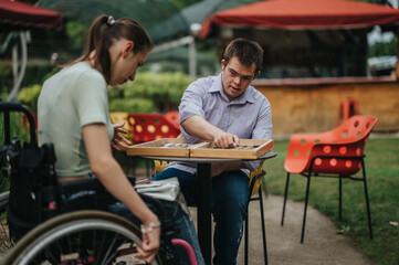 A boy with Down syndrome and a girl in a wheelchair enjoy playing backgammon together outside,...