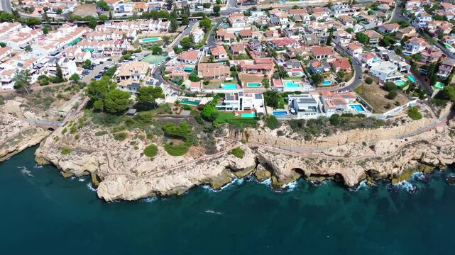 The drone flight along the rocky coastline of La Cala del Moral, Spain. Panoramic aerial view of the sea shore, residential sector, green gardens, pools. Sunny weather.