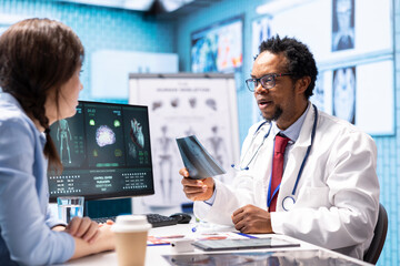 Professional medic analyzing x ray imaging and test findings in office, giving the diagnostic report for female patient at check up. African american specialist examines scans for interpretation.