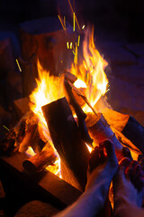 Tourist warming his feet by the fire at night. Flames and sparks fly upwards. Dark background, dramatic lighting. Outdoor recreation, travel, hiking and trekking in the mountains. Firewood is burning.