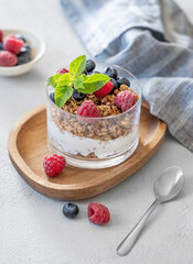 Natural yogurt with granola, raspberries and blueberries in a glass on a wooden board on a light background