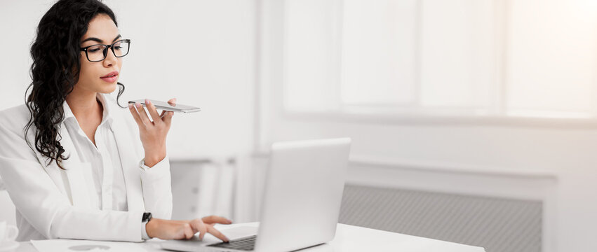 Virtual Assistant. Serious mexican businesswoman using mobile phone and working on laptop at modern office, copypace