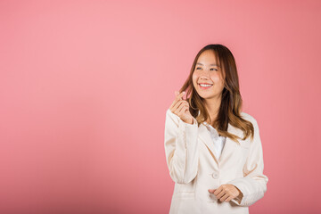 Confident business woman stands with a smile, creating a mini heart sign with her finger on a pink background. Asian portrait of a beautiful young female sending love for Valentine's Day, studio shot © sorapop