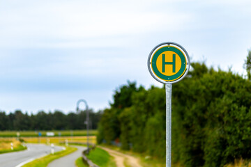 Bus stop sign located beside a winding road in a rural area with green fields under a cloudy sky
