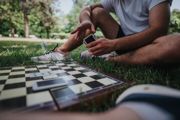 Fototapeta premium A young man sitting on the grass, playing cards next to a board game in a sunny park. The scene conveys relaxation and leisure amidst nature.