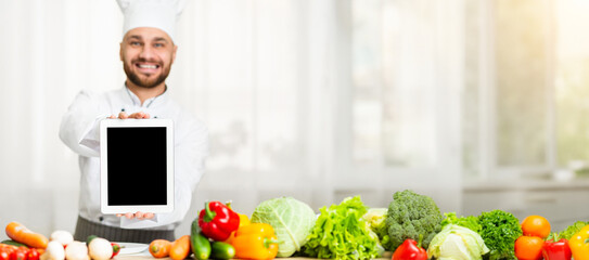 Joyful Chef Guy Showing Digital Tablet With Blank Screen To Camera Standing In Restaurant Kitchen Indoor. Shallow Depth, Mockup