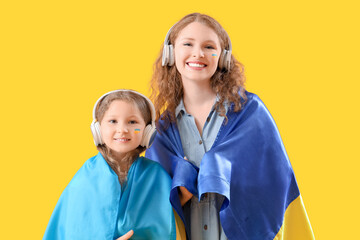 Happy little girl and her mother with Ukrainian flags listening music in headphones on yellow background