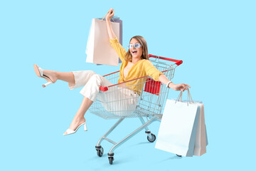 Stylish happy young woman with bags sitting in shopping cart on blue background