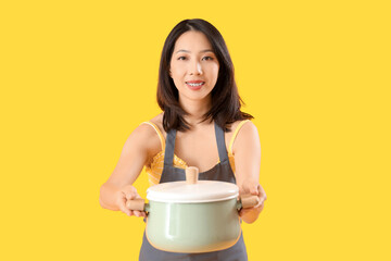 Young beautiful Asian woman with saucepan on yellow background