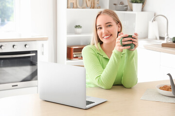 Young woman with cup of hot tea and laptop sitting at table in kitchen