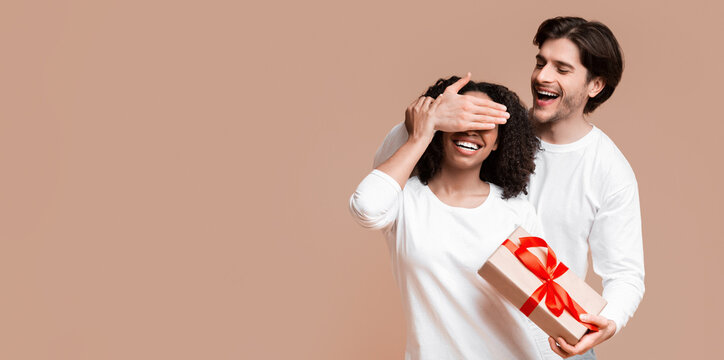 Gift For You. Loving Boyfriend Prepared Surprise To His Afro Girlfriend, Covering Her Eyes And Giving Present Box, Yellow Background With Free Space