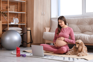 Beautiful sporty young pregnant woman with laptop and cocker spaniel practicing yoga at home