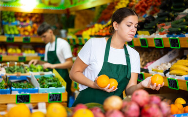 Hardworking young saleswoman working in a vegetable store puts fresh oranges on the counter