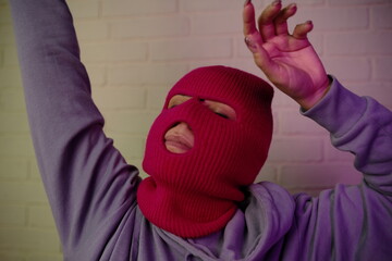 Individual in a pink balaclava poses against a colorful backdrop, making a gesture