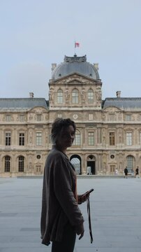 A woman with blue hair stands in front of a large building, holding a cell phone. The scene is quiet and peaceful, with no other people visible.