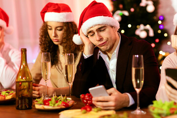 Bad Christmas Party. Bored Guy Using Smartphone During Dreadful Xmas Celebration With Friends At Home. Selective Focus