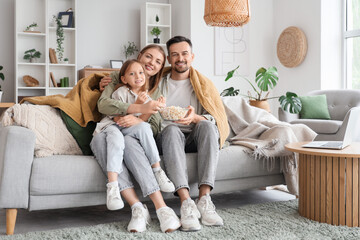 Little girl and her parents with warm plaid eating popcorn on sofa at home