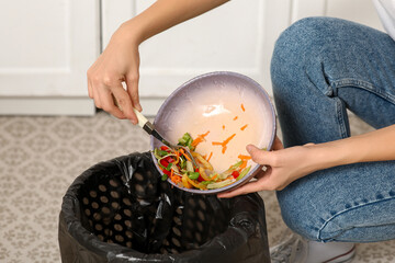 Young woman throwing organic food into trash bin in kitchen, closeup. Waste recycling concept