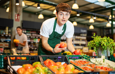 Cheerful young guy working in farm grocery store arranging ripe red tomatoes on counter with fresh...