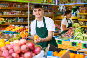 Positive man store employee displaying assortment of oranges at fruit department in supermarket