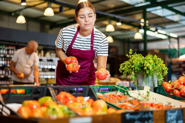 Skilled young female seller arranging organic ripe red tomatoes on vegetable counter at local farm...