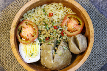 Meatball noodles are served in a round wooden bowl placed on a piece of burlap. top with chopped spring onions, sliced ​​red chilies and two slices of tomato.