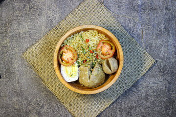 Meatball noodles are served in a round wooden bowl placed on a piece of burlap. top with chopped spring onions, sliced ​​red chilies and two slices of tomato.
