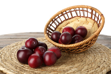 Fresh ripe plums and wicker bowl on wooden table against white background