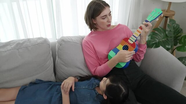 Asian cute daughter listening acoustic music while lie on her mother lap at sofa. Caucasian mother playing colorful ukulele while cute child looking at her parent. Family recreation concept. Pedagogy.