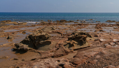 Close-up of eroded rocks forming intricate tide pools along a Mediterranean coastline, with the clear blue sea in the background under a bright sky, capturing natural coastal beauty