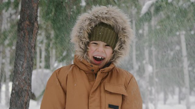 Medium close-up portrait of 7-year-old biracial boy in warm hooded parka, woolly hat standing under pine trees in winter forest on chilly day, having fun snow shower, looking at camera, laughing