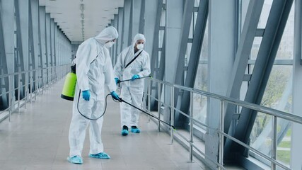 Two workers in protective gear spray disinfectant along a sleek, modern hallway. The environment is bright and spacious, highlighting the importance of hygiene during health crises.