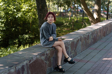 Fototapeta premium young girl with dyed hair, facial piercings and an oversize sweater holding a phone in her hand stares pensively into the distance while sitting in a summer park.