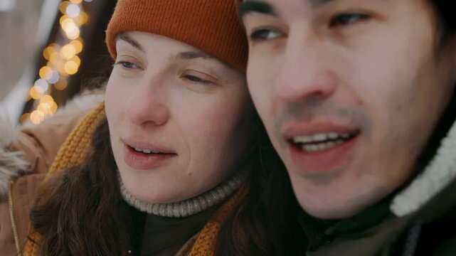 Close-up with bokeh lights of affectionate diverse young couple, Asian man and Caucasian woman in warm woolly hats and parka jackets sitting outdoors on cottage porch at Christmas, hugging, kissing