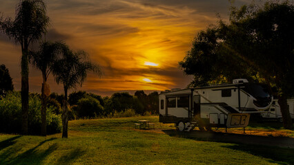 Rv motorhome fifth wheel parked at campsite under sunset sky with clouds and palm trees on grass