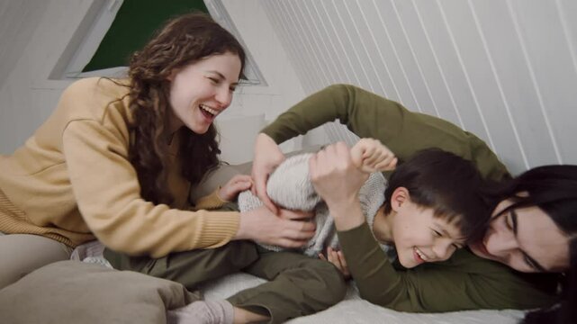 Full shot of happy young polyethnic family, Caucasian mom, Asian dad, son having playful fun scuffle together while relaxing on bed in attic, tickling each other, laughing and fighting with pillows