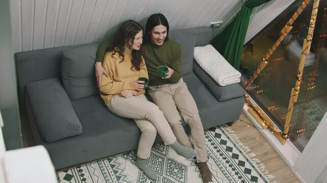 Full high-angle shot of Asian man and Caucasian woman sitting together on couch in holiday cabin, decorated with string lights, during romantic Christmas getaway, drinking coffee, hugging, chatting