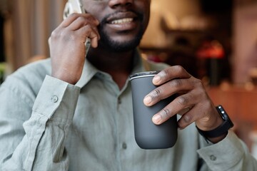Smiling man having conversation on phone while holding coffee cup in urban setting, wearing smartwatch and casual shirt. Indoor background suggesting cozy environment