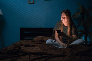 Smiling woman in pajamas sitting on bed read message on cell phone chatting online at night at...