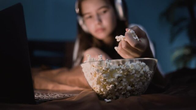 Close up of girl's hand taking popcorn from bowl while watching movie at night in bed