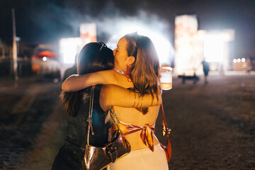 Two women embracing and kissing while enjoying a night at an outdoor music festival, with the...