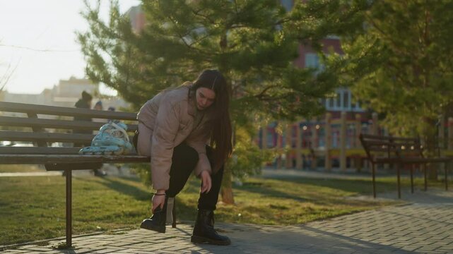 A girl in a peach jacket and black trousers is sitting on a bench in a park. She is seen taking off one boot while the rollerblades rest on the bench beside her