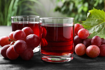 Tasty grape juice in glasses and berries on black wooden table against blurred green background,...