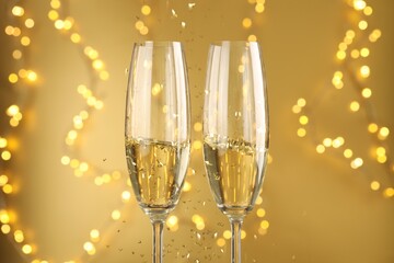 Glasses of sparkling wine and confetti against golden background with blurred lights, closeup. Christmas decor