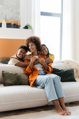 Funny African American children enjoying, embracing with their mother, sitting on sofa