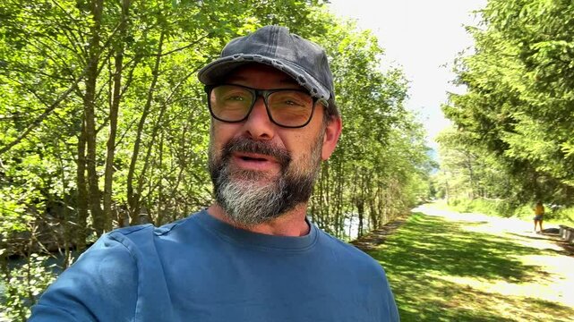 Adult male tourist in a blue shirt and cap video calling while hiking in the mountains, showcasing stunning panoramic views and sharing his travel adventure with social media followers